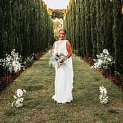 Frau in Brautkleid hält weißen Blumenstrauß mit grünen Blättern zwischen hohen Zypressen.