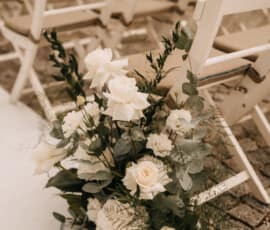 Weiße Blumendekoration mit Rosen und Eukalyptus an Stühlen einer Hochzeitszeremonie im Freien.
