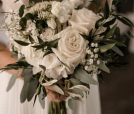 Braut hält einen weißen Brautstrauß mit Rosen, Schleierkraut und Eukalyptus vor ihrem Hochzeitskleid.