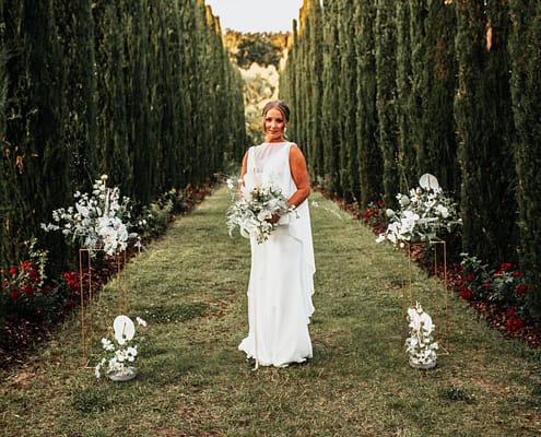 Frau in Brautkleid hält weißen Blumenstrauß mit grünen Blättern zwischen hohen Zypressen.