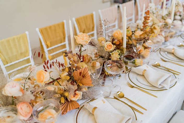 Tischdekoration mit rosa Rosen und Glasvasen | Gärtnerei Reinwald Tischdekoration mit rosa Rosen, Glasvasen, goldenen Bestecken und weißen Servietten auf einem langen Tisch.